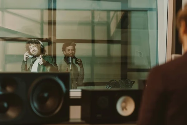 Jóvenes músicos felices grabando la canción detrás del vidrio en el estudio y mostrando los pulgares hacia arriba - foto de stock