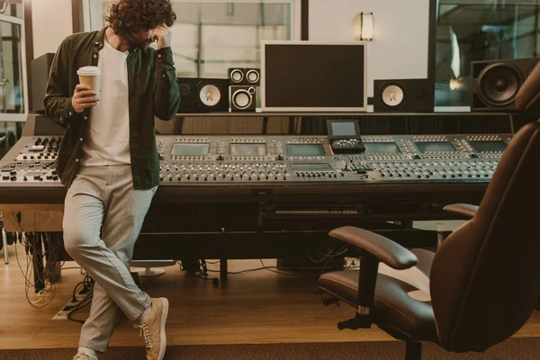 Joven productor de sonido de pie en el estudio con una taza de café para llevar - foto de stock