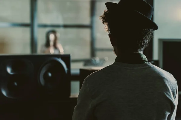 Vue arrière du producteur sonore regardant le chanteur enregistrer une chanson derrière un verre au studio — Photo de stock