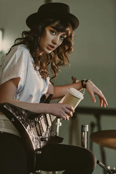 Bela músico feminino com guitarra elétrica e xícara de café para ir olhando para a câmera — Fotografia de Stock