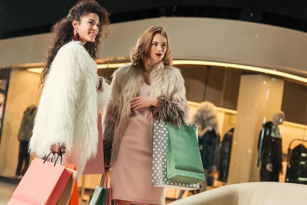 Hermosas mujeres jóvenes multiétnicas en abrigos de piel de moda sosteniendo bolsas de papel mientras compran juntos en el centro comercial - foto de stock