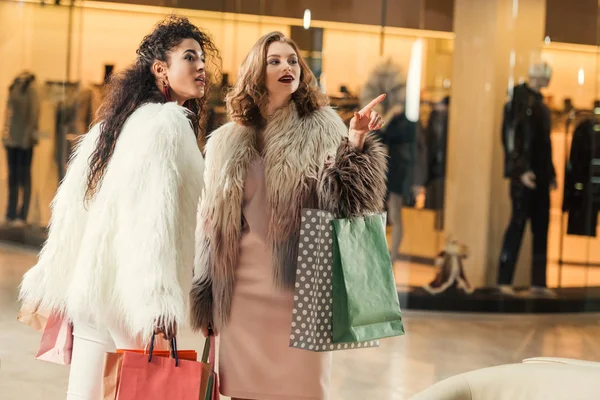 Mujeres multiétnicas de moda sosteniendo bolsas de papel y señalando con el dedo en el centro comercial - foto de stock