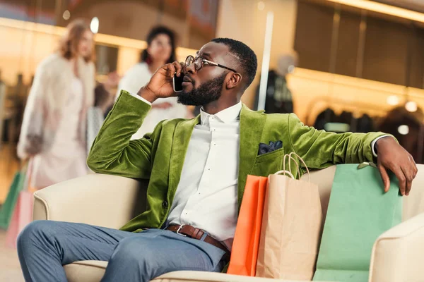 Guapo afroamericano hombre hablando por teléfono inteligente mientras está sentado con bolsas de compras en el centro comercial - foto de stock