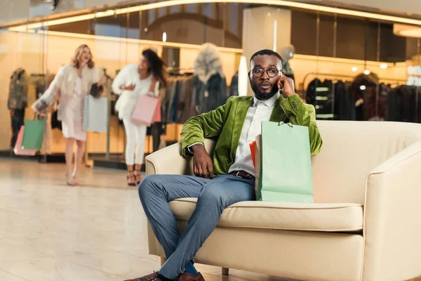Stilvoller afrikanisch-amerikanischer Mann spricht per Smartphone, während er mit Einkaufstüten in Einkaufszentrum sitzt — Stockfoto