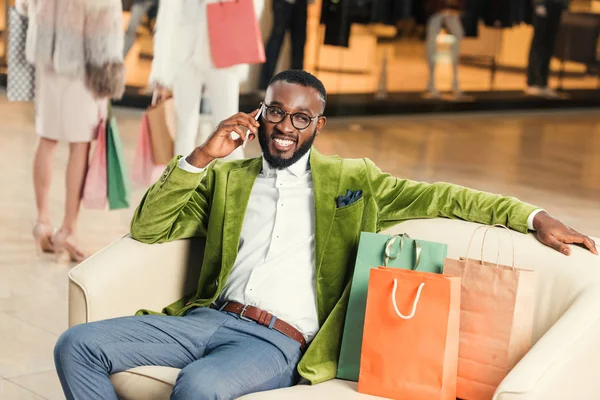 Guapo hombre afroamericano elegante hablando por teléfono inteligente mientras está sentado con bolsas de compras en el centro comercial - foto de stock