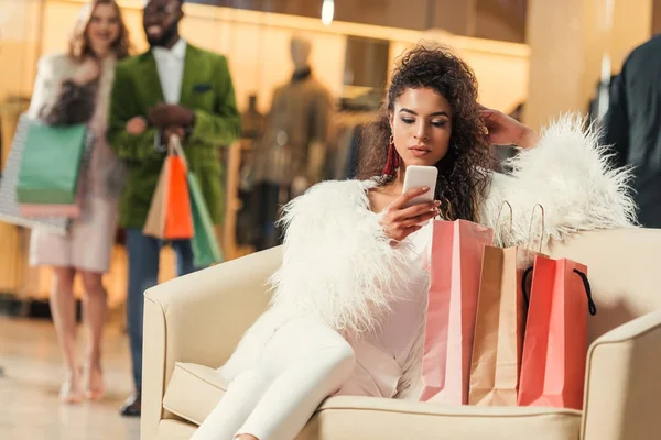 Jovem mulher americana africana moda usando smartphone enquanto sentado com sacos de compras no shopping — Fotografia de Stock