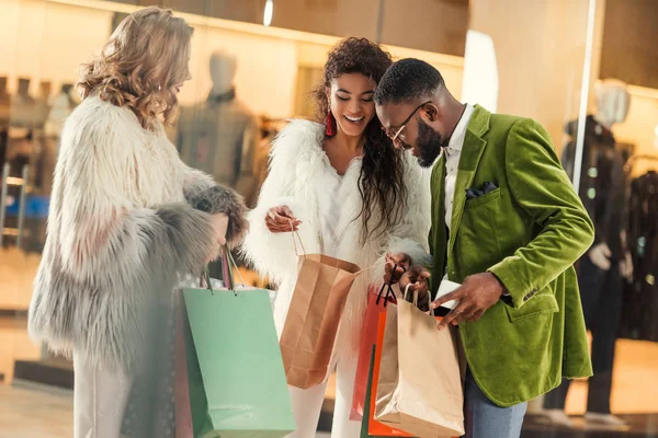 Glückliche stylische multiethnische Menschen, die in Einkaufszentren in Einkaufstüten schauen — Stockfoto