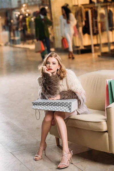 Nachdenkliche junge Frau mit Einkaufstasche sitzt auf Sessel und blickt in Einkaufszentrum in die Kamera — Stockfoto