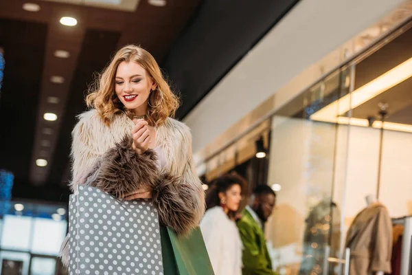 Smiling fashionable young woman in fur coat holding shopping bags while walking in mall — Stock Photo