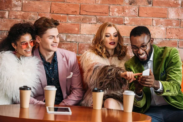 Smiling fashionable multiethnic young people using smartphone while drinking coffee — Stock Photo