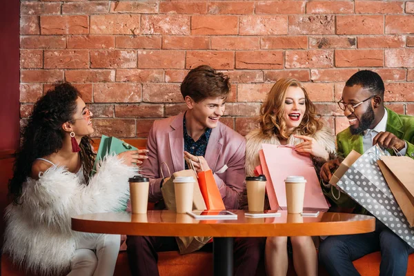 Стильные мультиэтнические молодые люди, глядя в сумки для покупок, выпивая кофе вместе — стоковое фото