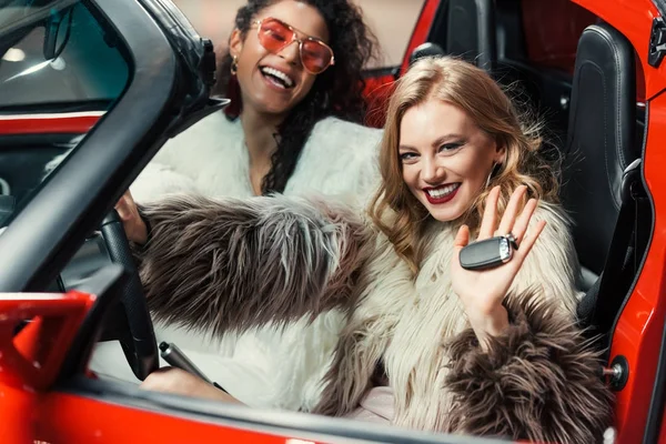 Счастливые стильные мультиэтнические женщины в шубах, держа ключи от машины, сидя в роскошном красном автомобиле — стоковое фото
