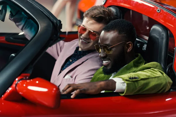 Счастливые стильные мультикультурные мужчины, сидящие в новом автомобиле в автосалоне — стоковое фото