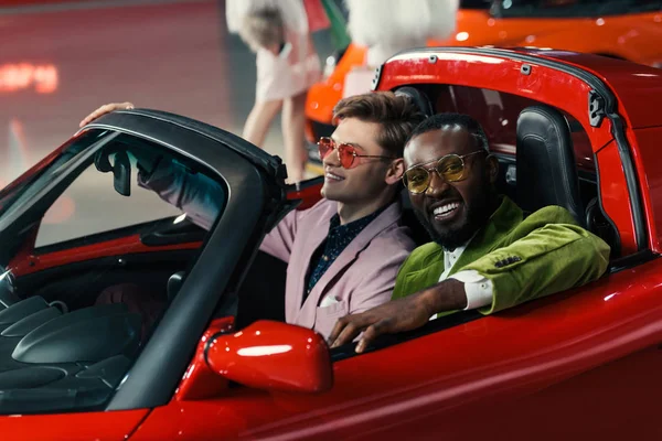 Улыбающиеся стильные мультиэтнические мужчины, сидящие в новом автомобиле в выставочном зале — стоковое фото