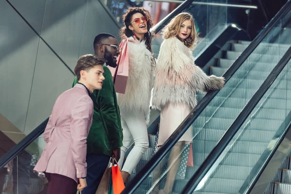 Stylish multiethnic young shoppers on escalator at mall with packages — Stock Photo