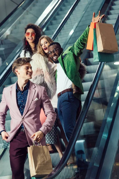 Felizes jovens compradores multiétnicos na escada rolante no shopping com pacotes — Fotografia de Stock