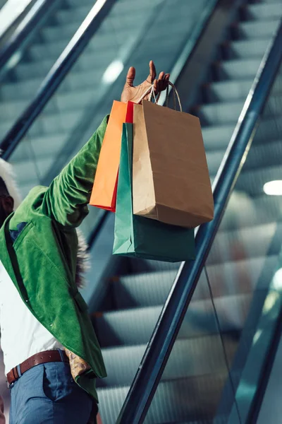 Recortado disparo de hombre sosteniendo bolsas de compras mientras monta escaleras mecánicas en el centro comercial - foto de stock