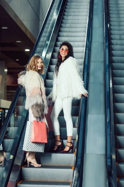 Mujeres jóvenes en abrigos de piel montando escaleras mecánicas en el centro comercial - foto de stock