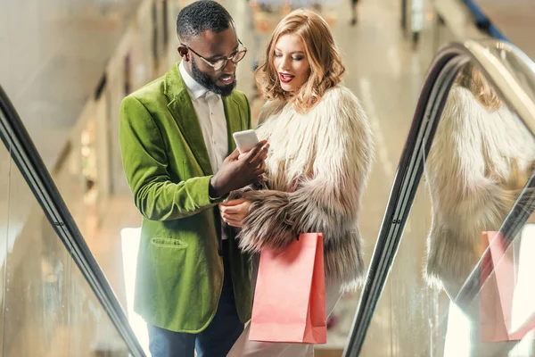 Pareja joven montando escaleras mecánicas y el uso de teléfonos inteligentes juntos en el centro comercial - foto de stock