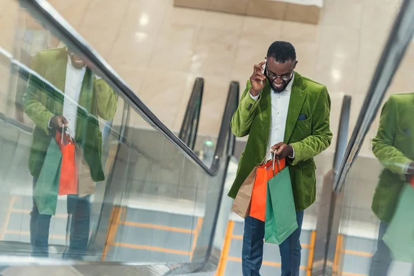 Sorrindo homem elegante com saco de compras falando por telefone na escada rolante no shopping — Fotografia de Stock