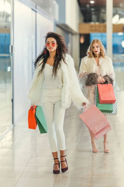 Femmes élégantes en manteaux de fourrure avec des sacs à provisions marchant par le centre commercial — Photo de stock