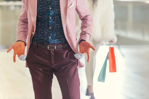 Cropped shot of man showing empty pockets while girlfriend holding shopping bags and walking away blurred on background — Stock Photo