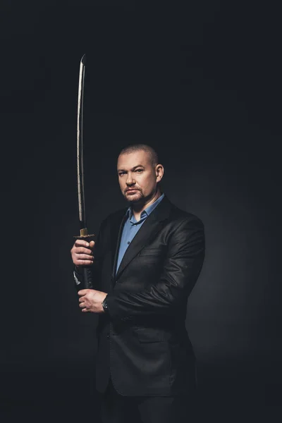 Samurai moderno en traje formal con espada katana mirando a la cámara en negro - foto de stock