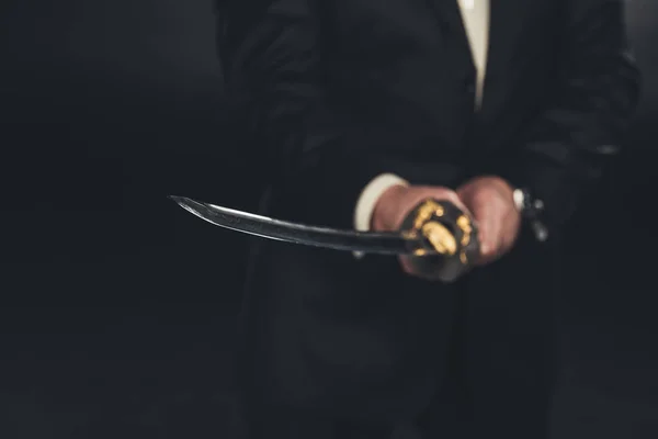 Recortado tiro de hombre en traje con espada katana sobre fondo oscuro - foto de stock