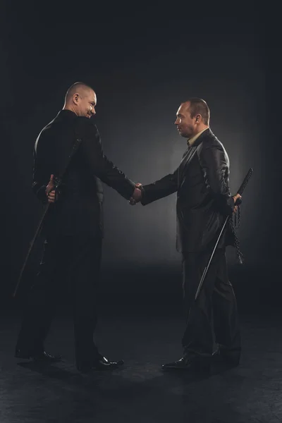 Side view of yakuza members shaking hands with katanas behind back isolated on black — Stock Photo