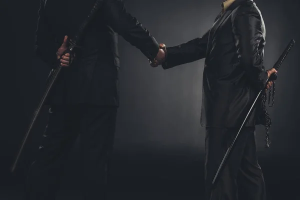 Cropped shot of yakuza members shaking hands with katanas behind back isolated on black — Stock Photo