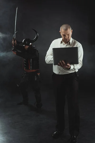 Hombre concentrado usando portátil mientras samurai de pie detrás de él con la espada - foto de stock