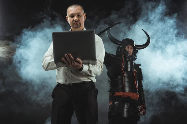 Hombre usando el ordenador portátil mientras samurai de pie detrás de él con espada - foto de stock