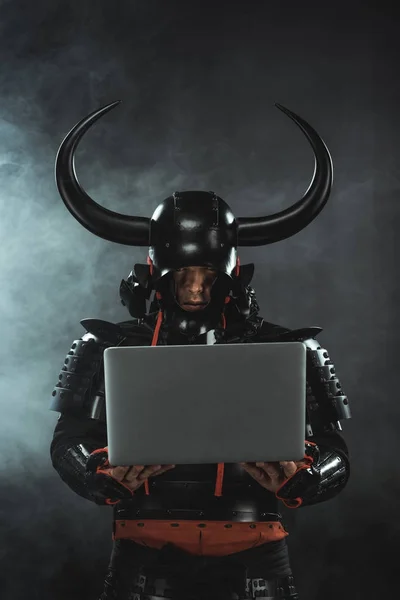 Armored samurai warrior using laptop on dark background with smoke — Stock Photo