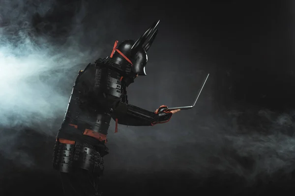 Seitenansicht von Samurai mit Laptop auf dunklem Hintergrund mit Rauch — Stock Photo