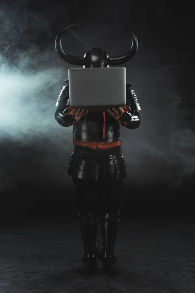 Armored samurai holding laptop on dark background with smoke — Stock Photo