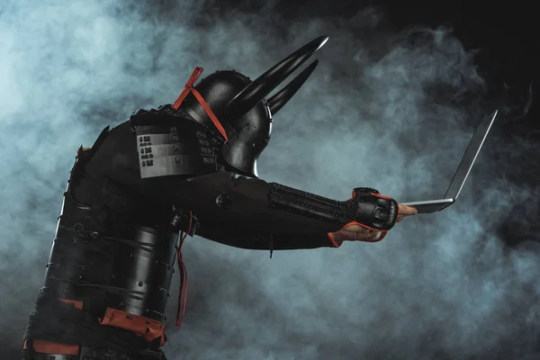 Вид збоку на броньований самурай, що тримає ноутбук на темному тлі з димом — стокове фото