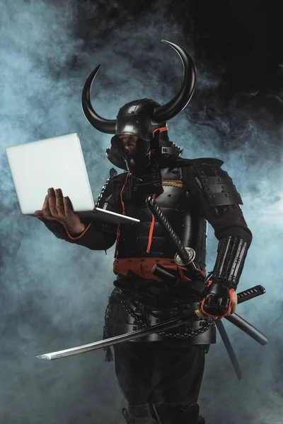 Samurai in traditional armor holding laptop on dark background with smoke — Stock Photo