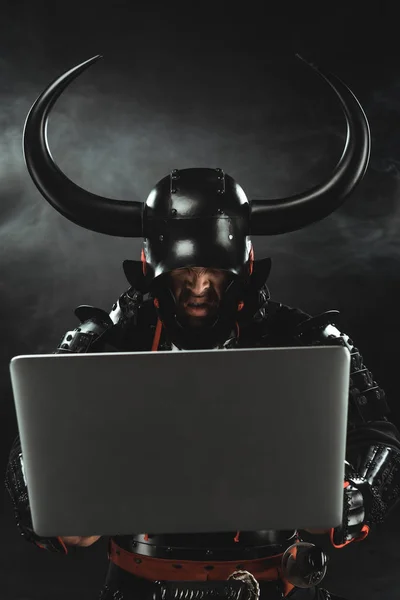 Samurai zangado em armadura usando laptop no fundo escuro com fumaça — Fotografia de Stock