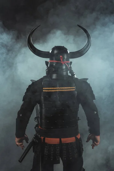 Vista trasera de samurai en armadura tradicional y casco con cuernos sobre fondo oscuro con humo - foto de stock