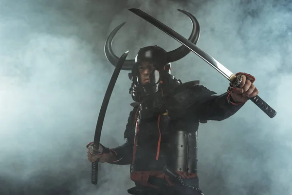 Samurai in traditional armor with dual katana swords in defence position on dark background with smoke — Stock Photo