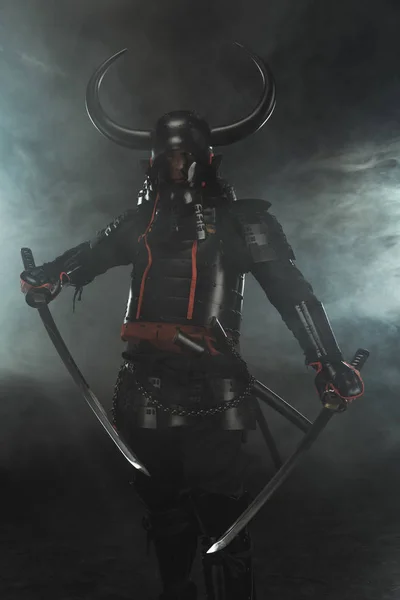 Samurai en armadura tradicional con espadas duales de katana sobre fondo oscuro con humo - foto de stock