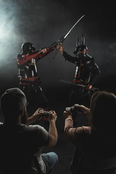 Back view of man and woman playing samurai fighting in real life with gamepads on black — Stock Photo