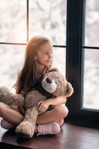 Linda criança sentada no peitoril da janela com ursinho de pelúcia — Fotografia de Stock