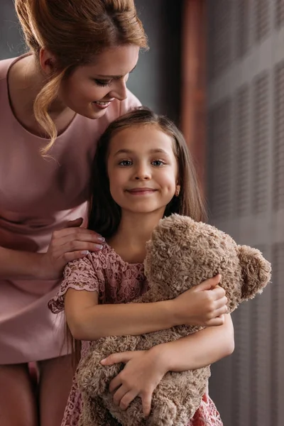 Young beautiful mother embracing her daughter while she holding teddy bear — Stock Photo