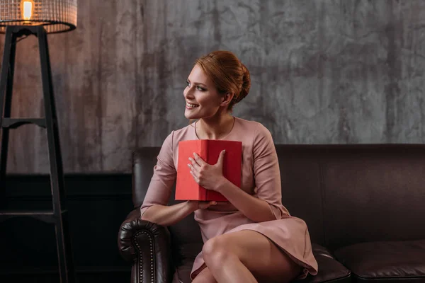 Feliz mujer joven pensativa con libro sentado en el sofá y mirando hacia otro lado - foto de stock