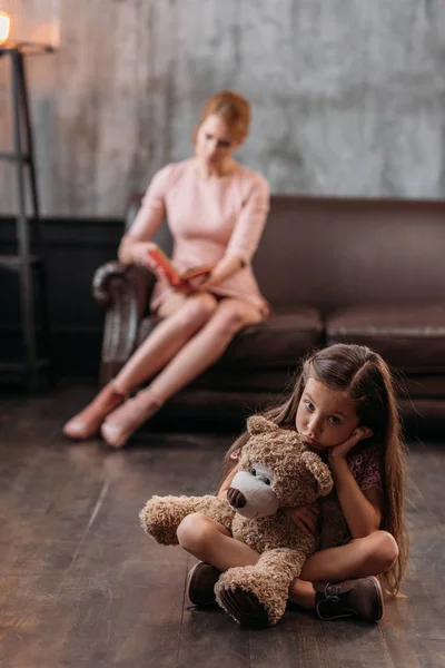 Pequena criança deprimida sentada no chão com ursinho de pelúcia, enquanto sua mãe sentada no sofá e livro de leitura — Fotografia de Stock