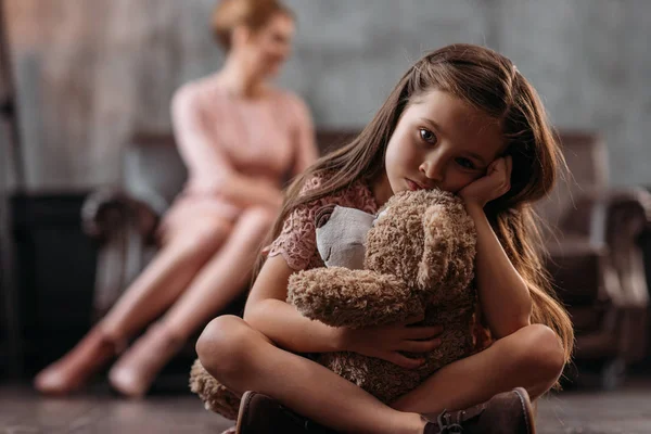 Pequena criança deprimida sentado no chão com ursinho de pelúcia, enquanto sua mãe sentada no sofá — Fotografia de Stock