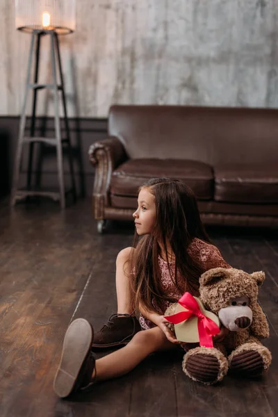 Adorável criança com caixa de presente em forma de coração e ursinho sentado no chão — Fotografia de Stock