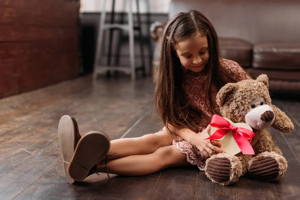 Glückliches kleines Kind übergibt Geschenkbox an Teddybär, während es auf dem Boden sitzt — Stockfoto