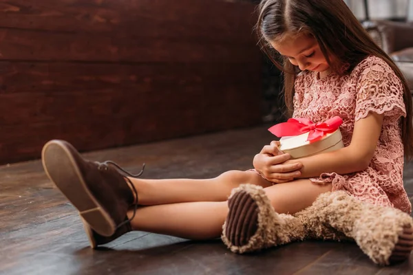 Entzückendes kleines Kind mit einer Geschenkbox in Herzform, während es auf dem Boden sitzt — Stockfoto